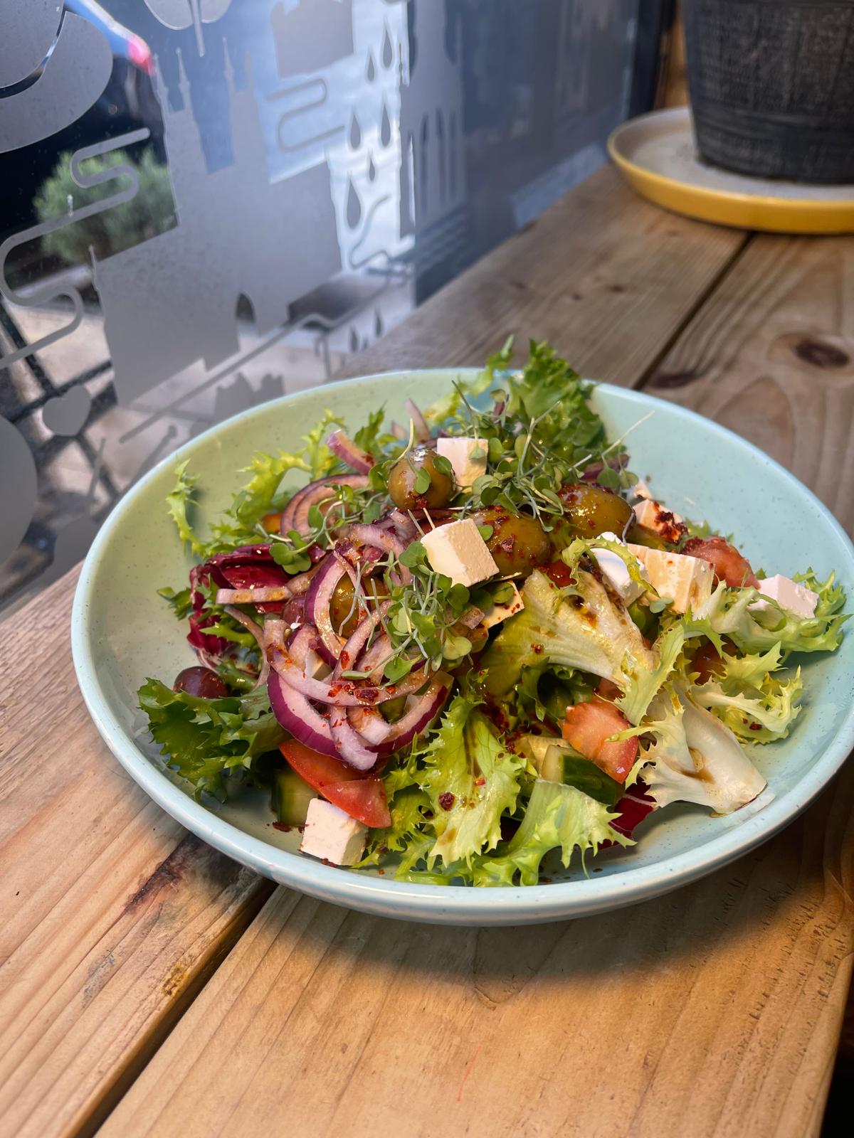 A salad in a pale green bowl: frisée, radicchio, red onion, tomato, cucumber, olives and cubes of feta with chilli flakes and a balsamic drizzle, by the window.