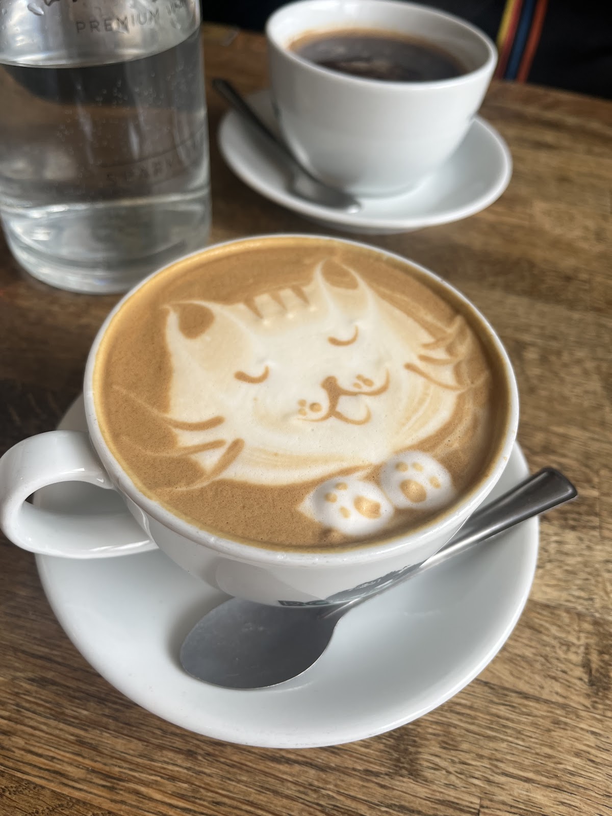 A latte with latte art of a sleeping cat with its paws on the rim, next to a black coffee and a bottle of water.