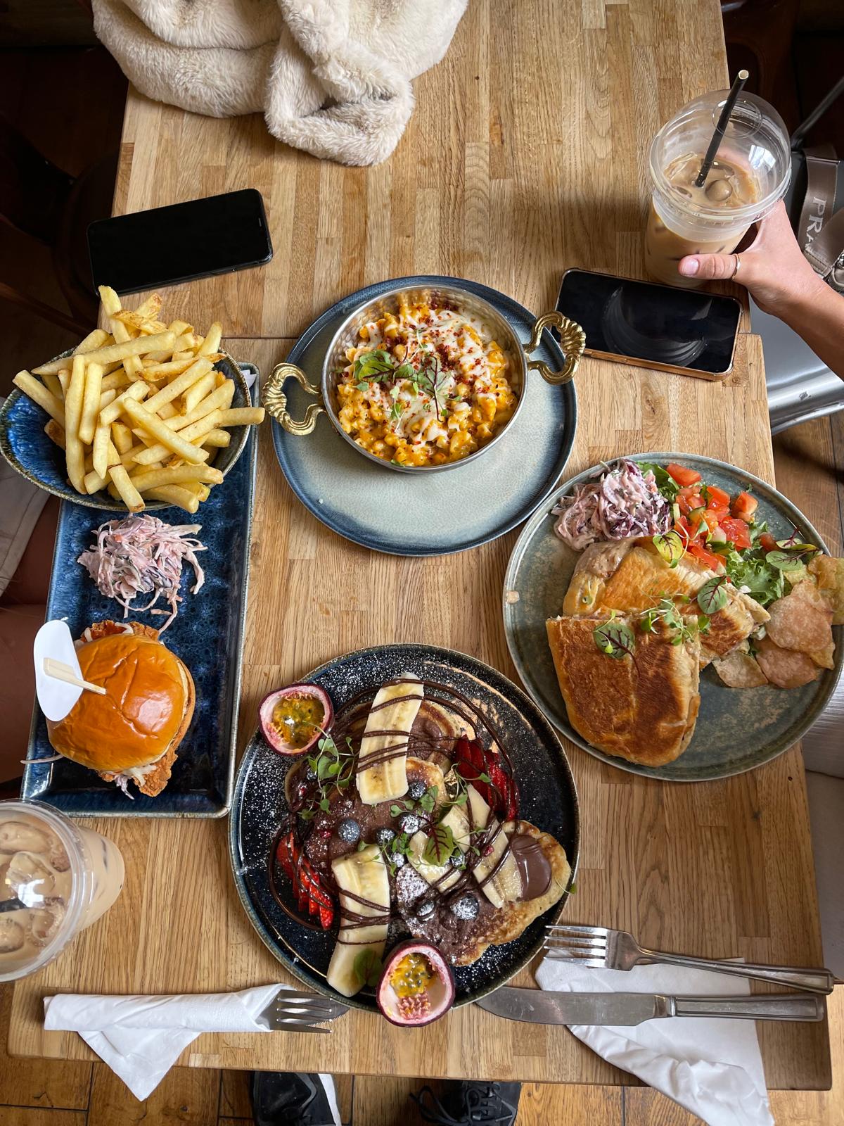 A table spread from above: pancakes with banana, berries, chocolate and passion fruit; a crispy chicken burger with fries and slaw; a toasted panini with salad; and a baked pasta dish in a brass-handled pan.