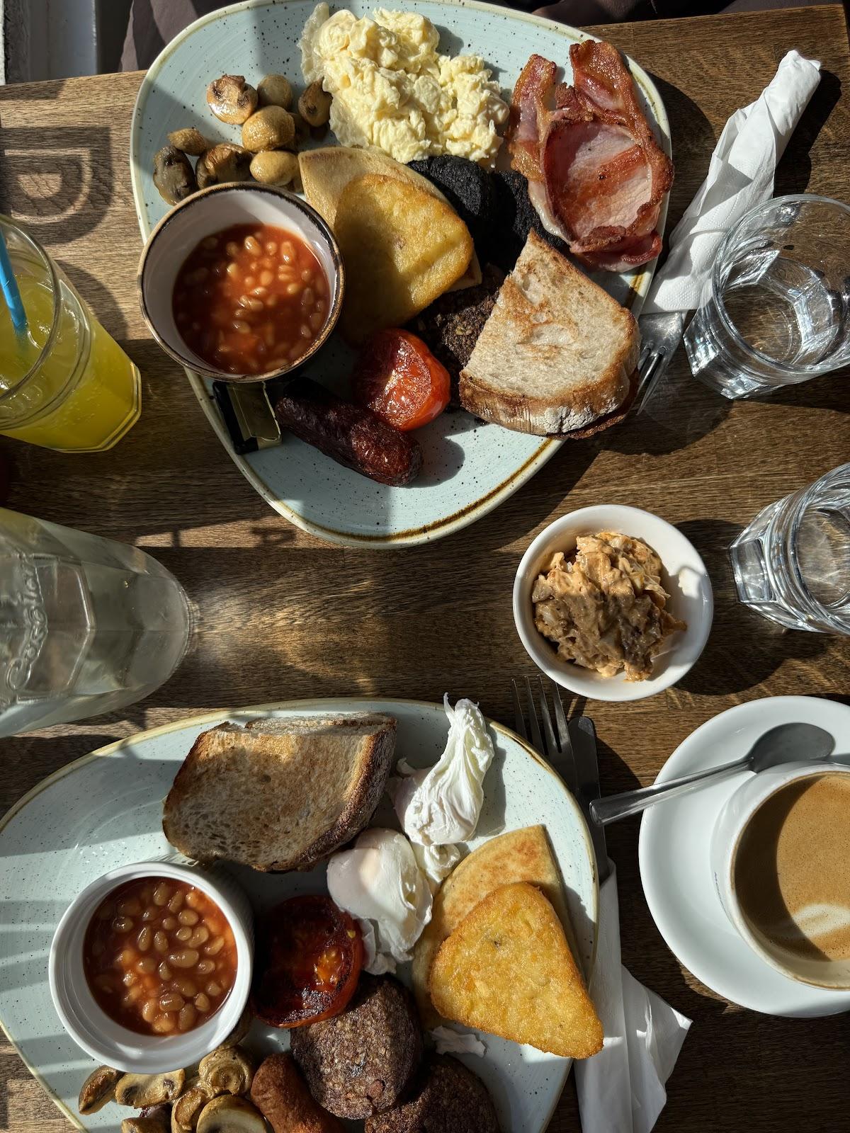 Two full breakfasts from above in morning sun: bacon, sausage, black pudding, haggis, tattie scone, hash brown, beans, mushrooms, tomato and eggs, with sourdough toast and coffee.