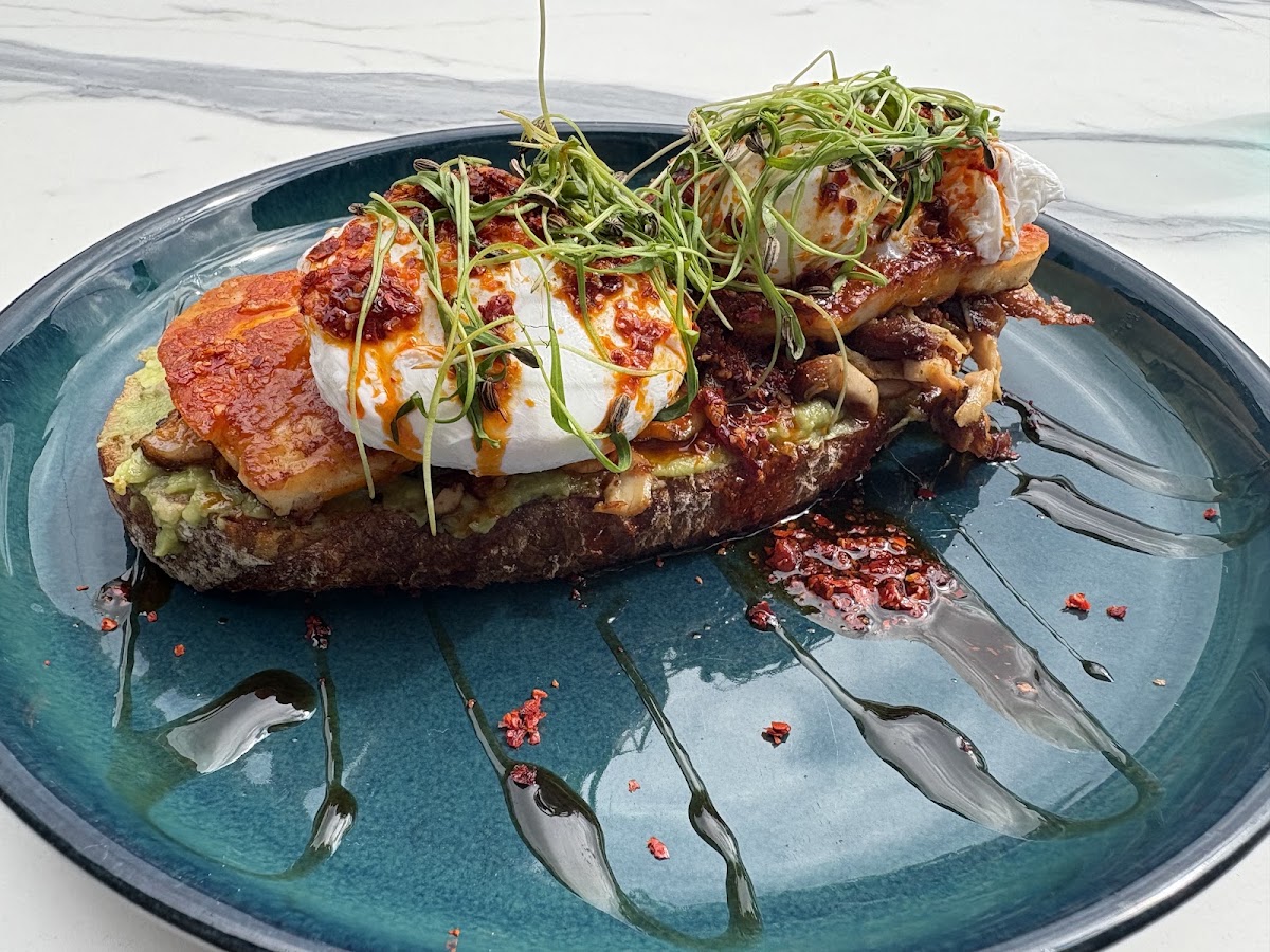 Two poached eggs on sourdough with avocado, grilled halloumi and mushrooms, under chilli butter and a tangle of pea shoots, on a deep teal plate.
